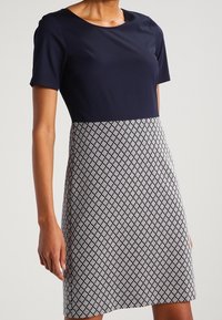 Kobieta w niebieskiej bluzce z krótkim rękawem oraz spódnicy do kolan w czarno-białym geometrycznym wzorze.