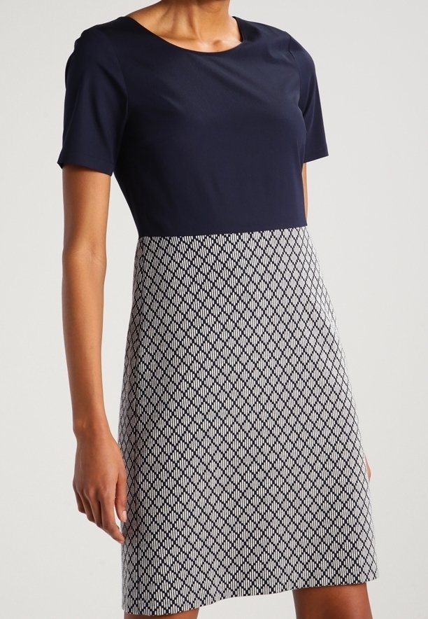 Kobieta w niebieskiej bluzce z krótkim rękawem oraz spódnicy do kolan w czarno-białym geometrycznym wzorze.