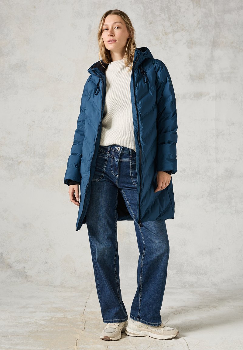 Doudoune bleue avec capuche, fermeture éclair et texture matelassée. Portée par-dessus un pull crème et un jean bleu foncé, assortie à des baskets claires.