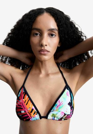 Frau mit lockigem Haar, die einen bunten Bikini-Top mit tropischem Muster und Neckholder trägt, die Hände hinter dem Kopf verschränkt vor einem weißen Hintergrund.