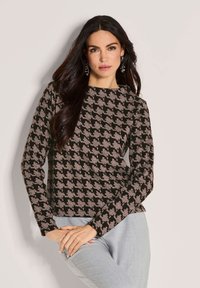 Vrouw met lang zwart haar in een zwart en beige hounds-tooth patroon langemouwtop en lichtgrijze broek, staande tegen een effen achtergrond.