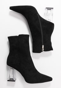 Botas de ante negras hasta el tobillo con puntas afiladas y cremallera lateral. Presentan un tacón bloque transparente con base negra. Textura suave.