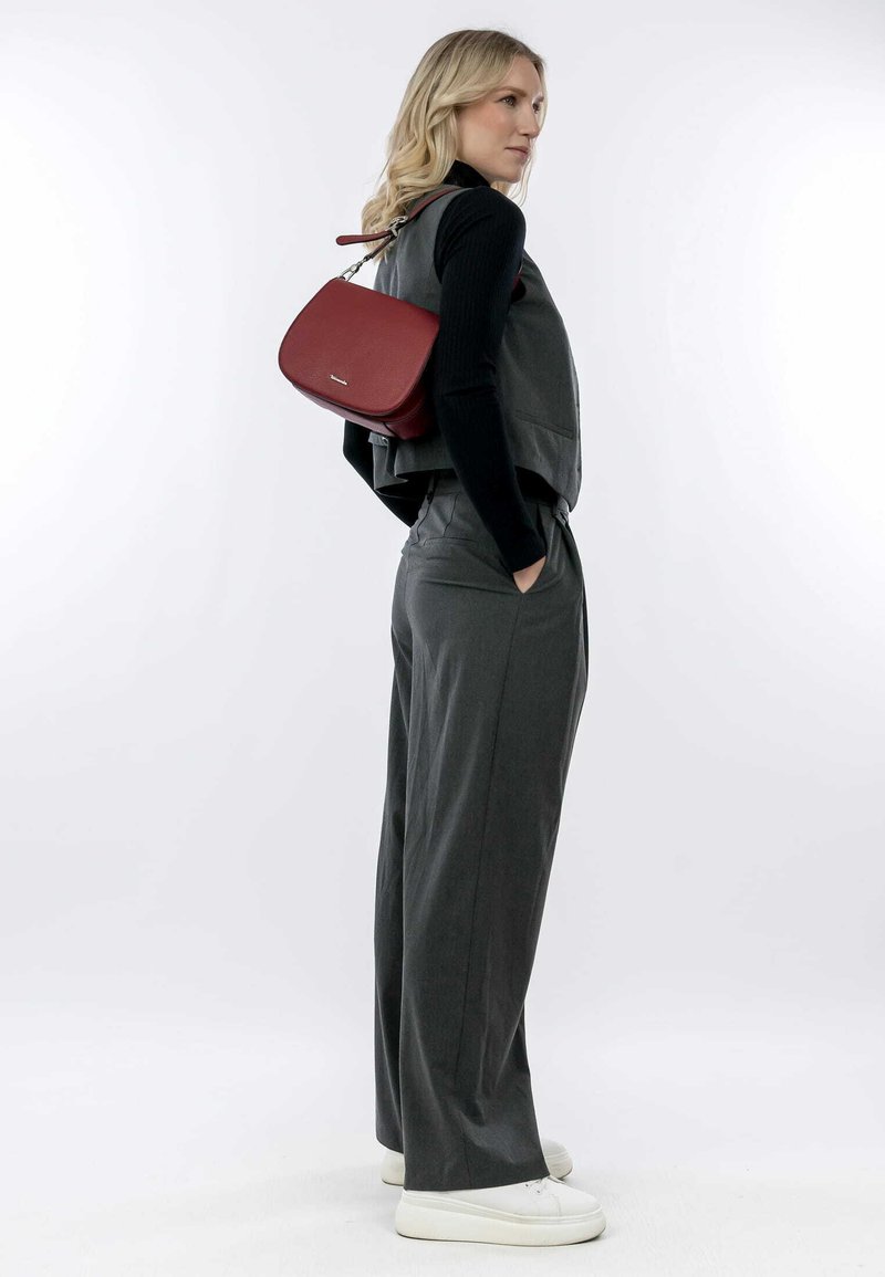 Chaleco gris ajustado y pantalones de pierna ancha, combinados con un bolso bandolera de cuero rojo. La modelo lleva zapatillas blancas y el cabello suelto. Fondo liso.