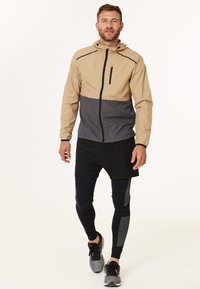 Beige und graue leichte Jacke mit Reißverschluss vorne, Kapuze und kleiner Brusttasche; getragen über schwarzen Shorts und Leggings; sportlicher Stil.