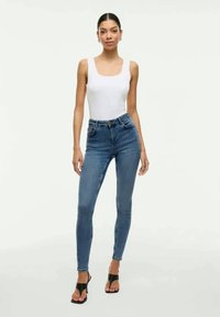 Calças de ganga azuis de cintura alta com um corte justo, feitas de denim, combinadas com uma blusa sem mangas branca e sandálias pretas de salto.