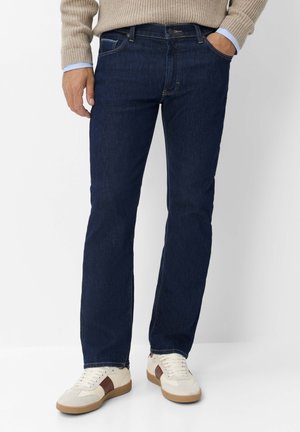 Mann trägt dunkelblaue Jeans, beigen Pullover mit blauen Hemdärmeln und weiße Sneakers mit braunen Streifen vor weißem Hintergrund.