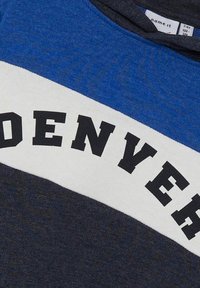 Sweatshirt bleu foncé et gris avec une large bande blanche portant le texte "DENVER" en lettres noires en gras sur la poitrine. Texture de tissu doux.