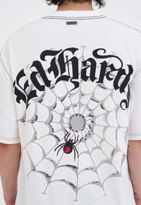 Person med en hvid skjorte med sort gotisk tekst "Ed Hardy" over et stort gråt spindelvæv og en sort edderkop med et rødt mærke.