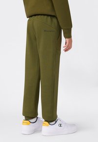 Pantalones de chándal verde oliva con cintura elástica, bolsillos laterales y un pequeño logo en azul marino en la parte trasera. Combinados con zapatillas blancas con acentos amarillos.