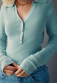 Haut en tricot côtelé bleu clair avec un col et une fermeture à six boutons. Manches longues avec trous pour les pouces. Associé à un bas en denim.