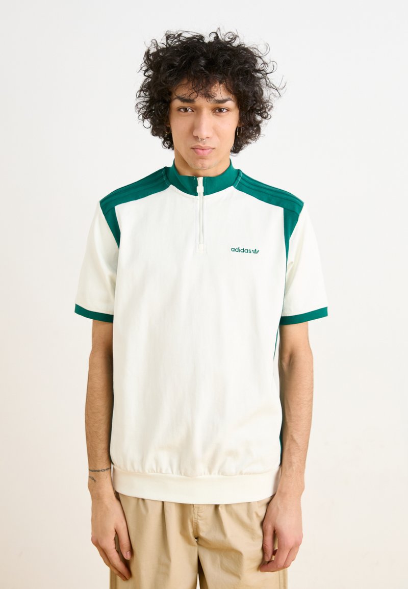 Λευκό πουλόβερ με πράσινες λεπτομέρειες, με ψηλό γιακά και κοντά μανίκια. Το ύφασμα φαίνεται μαλακό με κλείσιμο τύπου φερμουάρ στον λαιμό. Το λογότυπο της Adidas είναι εμφανές.