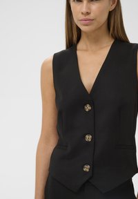 Juodas be rankovių liemenė su giliu V formos iškirpimu, penkiais bronziniais mygtukais ir šoninėmis kišenėmis, pagaminta iš lygaus audinio su tikslaus pritaikymo siluetu.