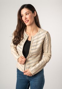 Giacca beige con zip e strisce metalliche diagonali, dotata di un collo rotondo e un design aderente. Indossata sopra una maglia nera, abbinata a jeans blu.
