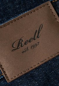 Brown leather label with embossed "Reell est. 1997" on dark blue denim fabric, featuring visible stitching and textured surface.