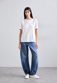 Tricou alb din bumbac, cu mâneci scurte și un mic logo auriu, purtat cu blugi albaștri, cu talie înaltă și lejeri, și adidași albi.