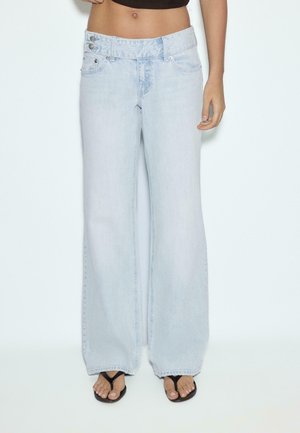 Person iført lyseblå vidde jeans og sorte flip-flops, stående med den ene hånd afslappet ved siden.