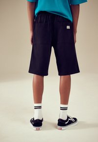 Shorts en denim noir à coupe décontractée, avec une taille élastique et deux poches arrière. Portés avec des baskets noires et des chaussettes blanches à rayures.