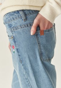 Jeans in denim azzurro con ricami di fiori sulla tasca, dotati di un'etichetta in pelle marrone e dettagli di cucitura visibili.