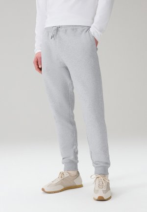 Mand iført lysegrå joggebukser med elastiske manchetter, hvide sneakers og en hvid langærmet skjorte, står med den ene hånd i lommen.