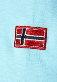 Hellesblaue Stoff mit einem rot-schwarz gestickten Patch der norwegischen Flagge. Die Nähte sind sichtbar und bieten texturreiche Details.