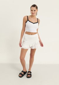 White ribbed crop top with black trim, paired with a white ruffled mini skirt. Black double-buckle sandals complete the outfit.