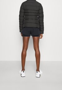 Černá péřová bunda s viditelným prošíváním, spárovaná s černými sportovními šortkami a bílými teniskami. Textura vypadá hladce a zatepleně.