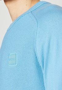 Gros plan d'une personne portant un pull bleu clair texturé avec un patch logo BOSS cousu sur la poitrine.