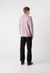 Polo majica s dugim rukavima, svijetlo ružičaste boje s visokim ovratnikom i bijelim detaljima, uparen s crnim hlačama i tamnim cipelama. Glatka tekstura.