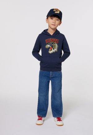 Hoodie marine avec un graphique de tigre et le texte "KENZO" ; associé avec un jean large bleu clair et des chaussures blanches avec des accents rouges ; casquette marine avec logo.