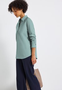 Camicia verde chiaro con colletto, maniche lunghe, polsini a bottone e orlo arrotondato, abbinata a pantaloni blu scuro e una borsa tote beige.