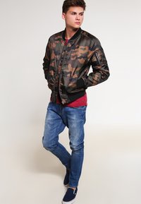 Karmas izgatavots bumbers ar kamuflāžas rakstu brūnās un zaļās nokrāsās, ar rāvējslēdzēju priekšpusē, ribota piedurkne un sānu kabata, saskaņots ar ziliem džinsiem un tumši zilām kurpēm.