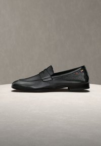 Mocassin en cuir noir pour homme avec un petit talon et un petit détail en tissu coloré près du talon, placé sur une surface claire.