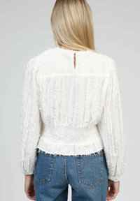 Femme blonde portant un blouse longue à manches longues blanche texturée avec taille smockée et jean en denim bleu, vue de dos.