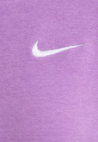 Bílá výšivka loga Nike swoosh na pozadí pletené látky v levandulové barvě.