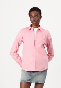 Camicia rosa a maniche lunghe con colletto button-down, caratterizzata da una vestibilità slim e una tasca con dettaglio logo. Indossata sopra una camicia bianca.