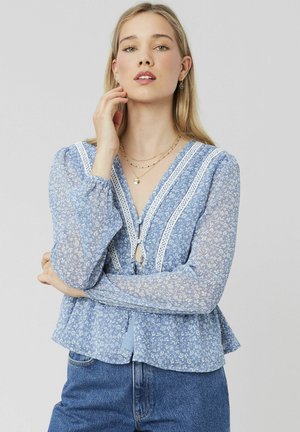 Blouse bleu clair avec motif floral, accents en dentelle, décolleté en V, manches longues et transparentes, et un bas en peplum, associée à un jean en denim bleu.