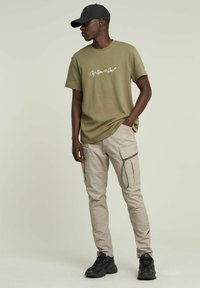 T-shirt in cotone verde oliva con logo in bianco, abbinata a pantaloni cargo beige chiaro con tasche con zip e sneakers nere.