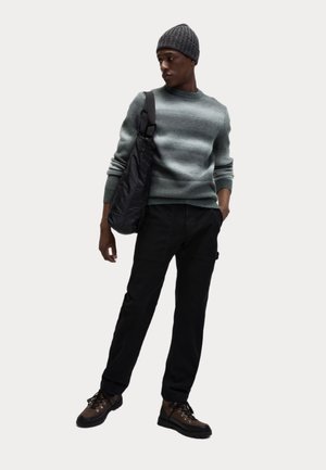 Suéter gris de punto, pantalones cargo negros, botas de senderismo marrones y un gorro gris; el modelo sostiene un bolso de hombro negro en una pose de pie.