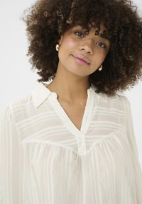 Blusa leggera e trasparente color crema con righe verticali, scollatura a V e un sottile design plissettato. Orecchini rotondi in oro completano il look.