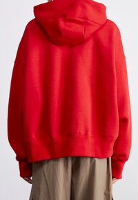 Röd oversized hoodie med dragsnodd i huvan, ribbade ärmslut och fåll. Tillverkad av mjuk bomullsblandning med en slät textur. Visad bakifrån.