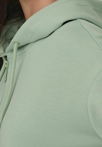 Světle zelená zipová mikina s kapucí, která má měkkou texturu, stahovací šňůrku a šikmé švy pro uvolněný střih.