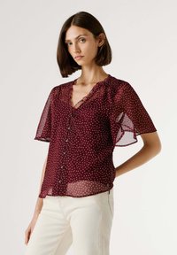 Blouse transparente bordeaux à pois blancs, col en V, fermeture à boutons, manches courtes évasées et détail de col froncé.