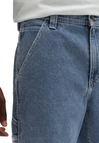 Vaaleansiniset farkut, joissa on klassinen viiden taskun muotoilu, vetoketjukiinnitys ja hienovaraista kulumista kankaassa. Teksturoitu puuvillamateriaali.