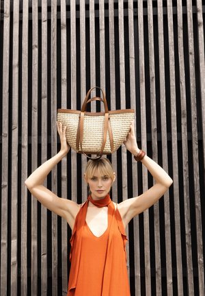 Woman in orange sleeveless dress holding a woven handbag with brown leather straps above her head against vertical wooden slats background.