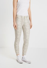 Jeans ajustados de estampado de serpiente en color beige claro con acabado brillante, con una cintura de altura media y cinco bolsillos. Combinados con zapatillas altas blancas.