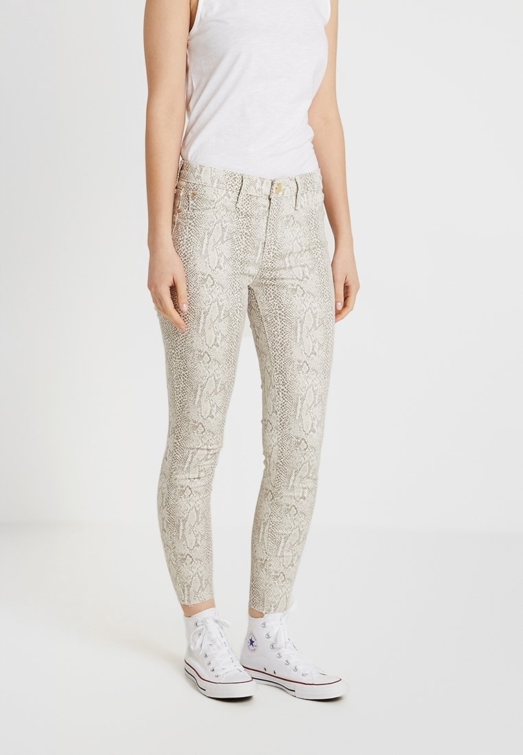 Jeans ajustados de estampado de serpiente en color beige claro con acabado brillante, con una cintura de altura media y cinco bolsillos. Combinados con zapatillas altas blancas.