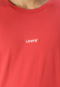 T-shirt roșu din bumbac, cu guler rotund, mâneci scurte și etichetă LEVI'S albă cusută pe pieptul stâng. Textură netedă, design casual.