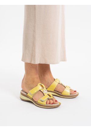 Yellow patent sandals with two wide straps, a circular decorative button, and a textured rubber sole. Worn with a ribbed beige skirt.