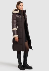 Rjava oblazinjena jakna s kapuco, z kremasto barvo umetnega krzna na robovih, žepki z zadrgo in ozko silhueto. Nošena s črnimi škornji do kolen.