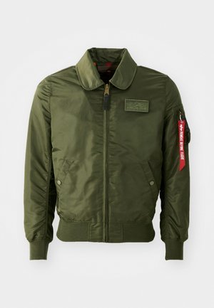 Olívazöld bomber dzseki cipzáras lezárással, kerek nyakkal, bordázott mandzsettákkal, két oldalzsebbel és piros szövet címkével az ujjon.